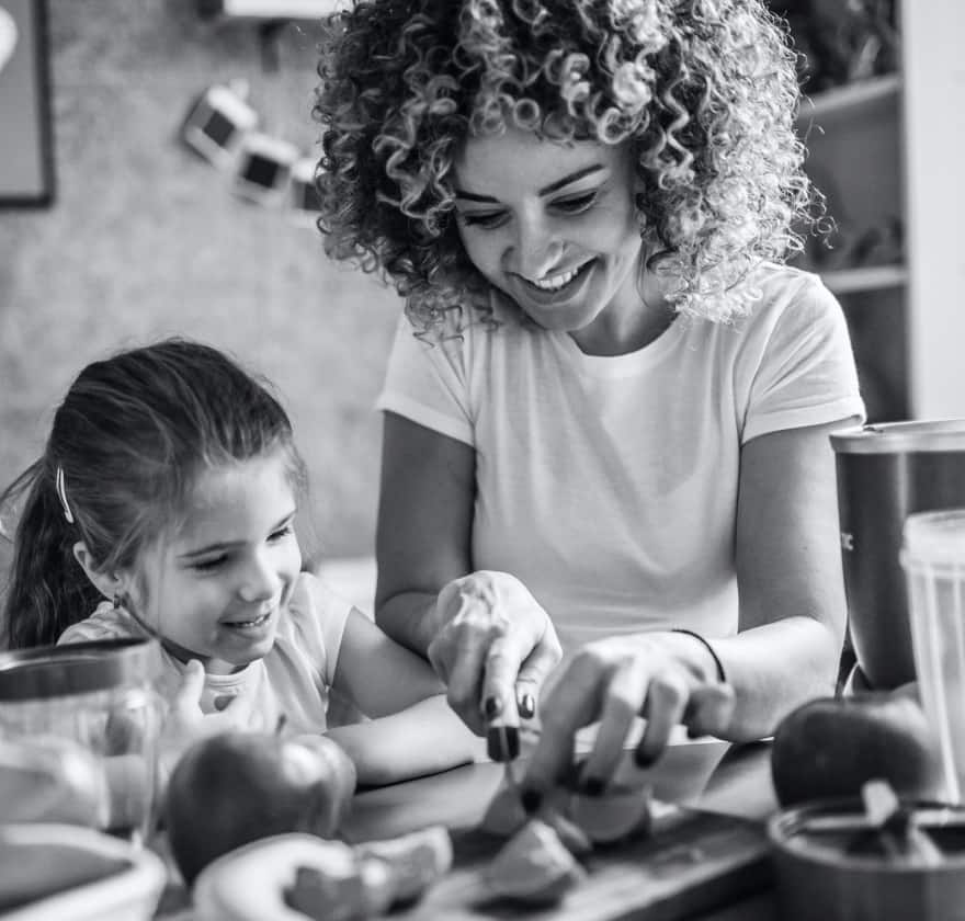 Black and white photo of a woman standing next to a young girl cutting fruits on a cutting board next to a nutribullet® pro blender